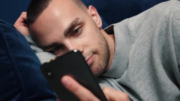 Молодий какасієць користується смартфоном, сидячи на дивані вдома. Man Browses через Інтернет, переглядає відео та використовує соціальні мережі вдома. — стокове відео