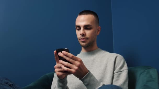 若い白人男性は自宅で椅子に座っている間にスマートフォンを使用します。インターネットを通して男が閲覧し、ビデオを見て、自宅でソーシャルネットワークを使用しています. — ストック動画