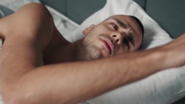 Close up shot of sleepy Caucasian man waking up in the morning, taking a smartphone from the bedside table and turning off its alarm clock to get more sleep
