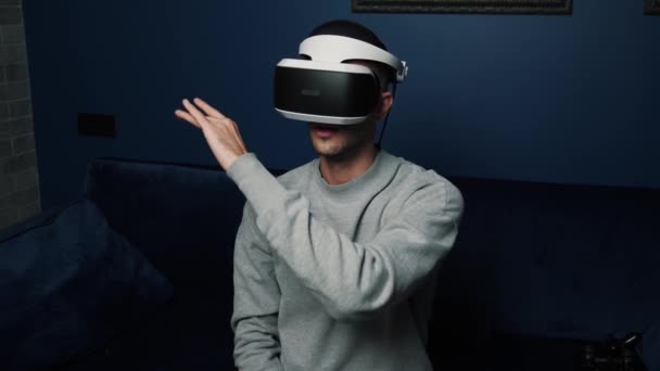Caucasian young man taking on VR headset, scrolling and taping in the air while sitting on the sofa at a home. A handsome beautiful man wearing a virtual reality headset and scrolling virtual display — Stock video