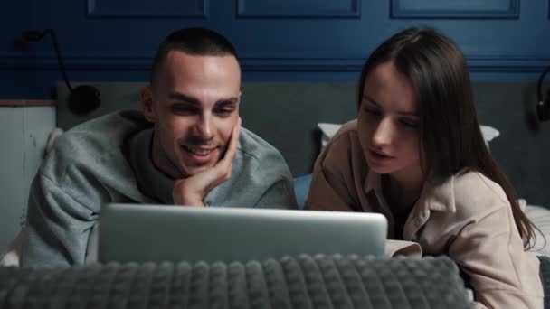 Glückliche Mann und Frau mit Laptop Computer zu Hause beobachten Online-Show zusammen im Bett liegen. Junges erwachsenes Paar schaut auf Laptop und macht Videoanruf im Schlafzimmer zu Hause. — Stockvideo