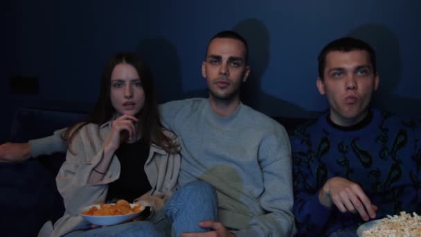 Los jóvenes se relajan en el sofá de casa mientras ven una película de terror y comen palomitas de maíz. Amigos asustados de ver una película de suspenso en la televisión moderna en la sala de estar. — Vídeos de Stock