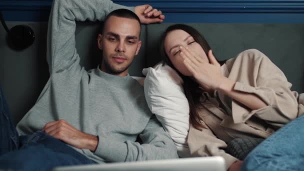 Casal cansado relaxando em um sofá e assistindo a um filme no laptop em casa. Retrato de um homem caucasiano cansado e mulher caucasiana em um sofá bocejando assistindo filme em casa — Vídeo de Stock