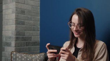 Kumar oynayan genç bir kadın evde cep telefonuyla oyun oynuyor. Çekici, gülümseyen bir kadın akıllı telefon oyunlarından zevk alıyor. Evdeki koltukta oturuyor..