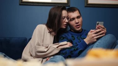 Couple relaxes on couch at home, caucasian man showing on smartphone to woman videos, photo, take selfie family picture, make video call having fun.