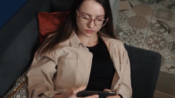 Hazard młoda kobieta grać w gry na telefon komórkowy w domu. Atrakcyjna kobieta ciesząca się grami wideo na smartfonie siedząc na sofie w domu. — Wideo stockowe