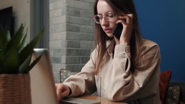 Молода білошкіра бізнес-леді професійно розмовляє по телефону, використовуючи ноутбук, сидячи на домашньому офісному столі . — стокове відео