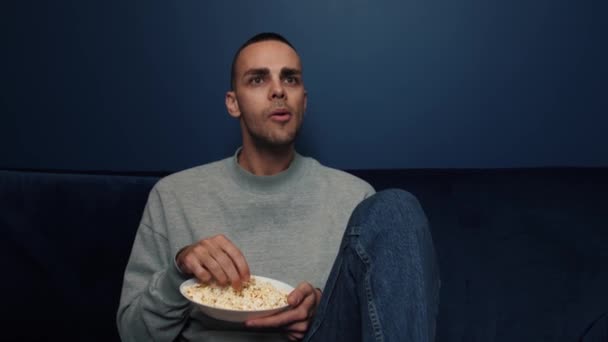 Zájem, vášeň, nadšení. Muž sám v pokoji se večer dívá na televizi a jí popcorn, velmi se zajímá o to, co vidí na plátně a nemůže odtrhnout oči. Detailní záběr — Stock video