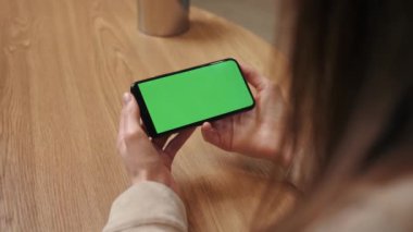 Woman hands holding smartphone horizontal mobile device with green display in home interior - over shoulder close up view. Mock up, chroma key, template, green screen, technology concept