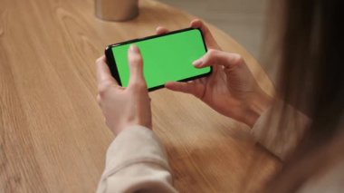 Woman hands holding smartphone horizontal mobile device with green display in home interior - over shoulder close up view. Mock up, chroma key, template, green screen, technology concept