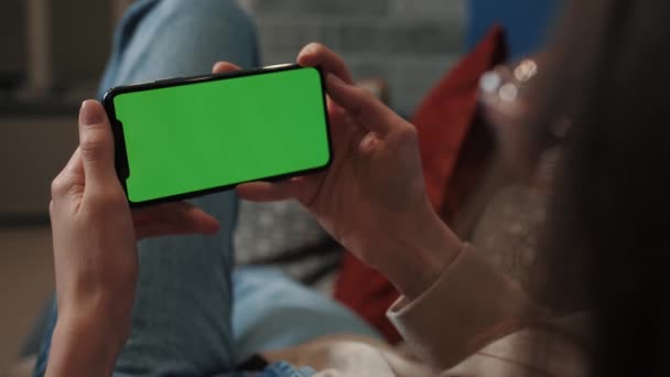 Woman hands holding smartphone horizontal mobile device with green display in home interior - over shoulder close up view. Mock up, chroma key, template, green screen, technology concept — 图库视频影像
