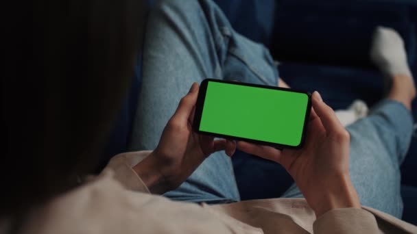 Woman hands holding smartphone horizontal mobile device with green display in home interior - over shoulder close up view. Mock up, chroma key, template, green screen, technology concept — Wideo stockowe