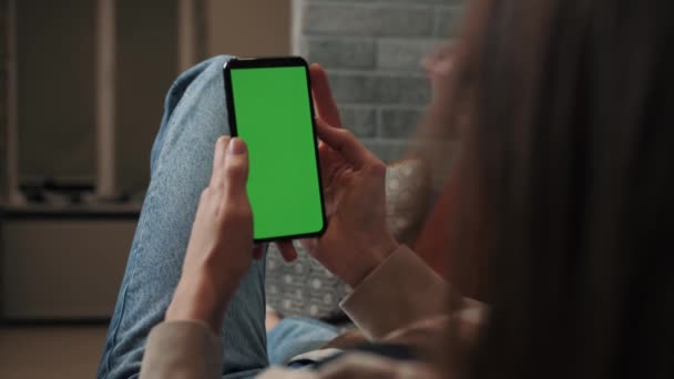 Woman hands holding smartphone mobile device with green display in home interior - over shoulder close up view. Mock up, chroma key, template, green screen, technology concept — ストック動画