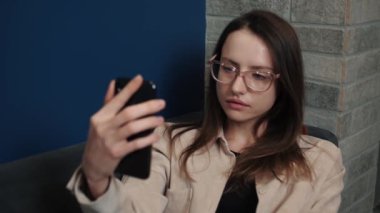 Kafkasyalı iş kadını dahili ofiste görüntülü konuşma yaparak akıllı telefondan konuşuyor. Ev içi ofis içi görüntülü görüşme için telefon kullanan bir kadın. İş kadını cep telefonu kamerasına el hareketi yapıyor..