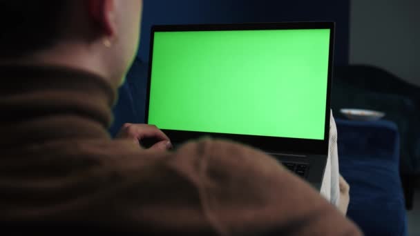 Over skulderbildet av en mann som jobber med en bærbar PC på sofaen hjemme med grønn skjerm, kromatøkkel, gjør videosamtaler med venner, familie, jobber med en bærbar PC på hjemmekontoret. – stockvideo