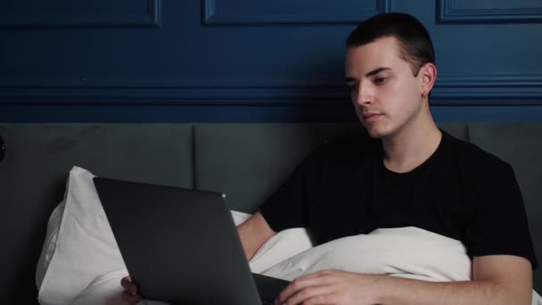 Müder junger kaukasischer Mann, der spät in der Nacht am Laptop auf dem Bett arbeitet und tippt, dann gähnt, als ob er einschläft. — Stockvideo