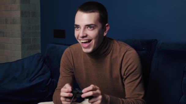 Счастливый взволнованный молодой кавказский мужчина геймер держит контроллер, играющий в видеоигры, сидя на диване, чувствуя себя сверх радости в видеоигре в одиночестве дома — стоковое видео