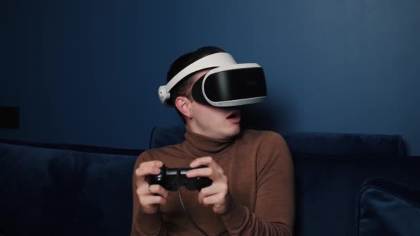 Atractivo hombre caucásico mantenga el joystick y use casco de gafas de realidad virtual en el sofá en casa. Gamer juego usando gafas VR en casa. — Vídeo de stock