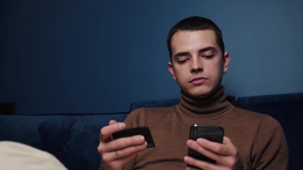 Молодий білий чоловік тримає кредитну картку і смартфон, сидячи на дивані вдома. Чоловік покупця використовує миттєві легкі мобільні платежі, роблячи покупки в інтернет-магазині. Служба додатків електронного банкінгу — стокове відео