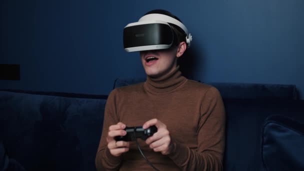 Un homme caucasien attrayant tient un joystick et porte un casque de lunettes de réalité virtuelle sur un canapé à la maison. Bonheur gamer jouer à l'aide de lunettes VR à la maison. — Video