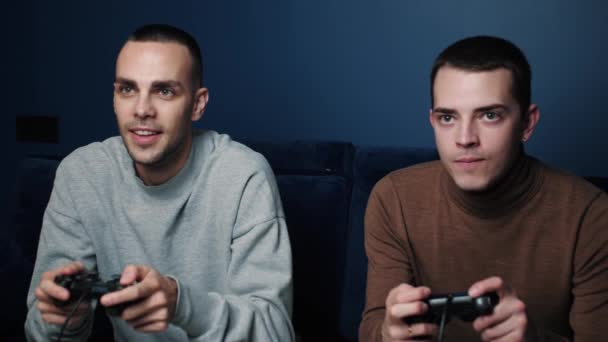 Amigos sonrientes con mandos joystick controlador jugando videojuegos en el sofá en casa. Retrato de chicos caucásicos jugando con la consola relajándose en el sofá en casa. — Vídeos de Stock