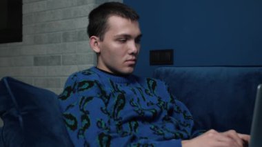 Rahatlamış bir adam ev ofisindeki kanepede dizüstü bilgisayarı kullanarak esniyor, bilgisayarını dairede uzaktan daktilo etmeye odaklanıyor.