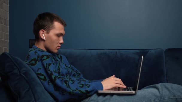 Relaxed man student freelancer met behulp van oortelefoons laptop apparaat leunend op de bank thuis kantoor, gericht werken op afstand typen op laptop in appartement — Stockvideo