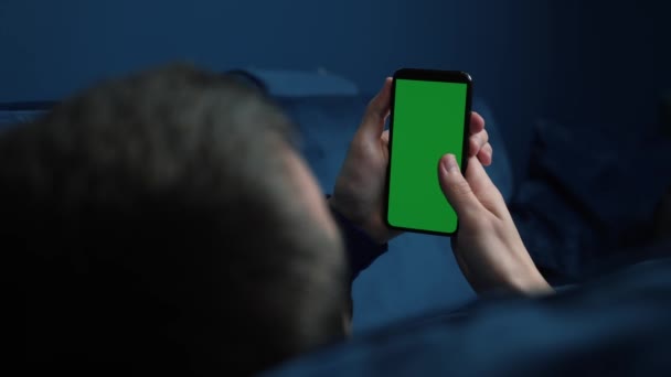 Hombre acostado en el sofá usando un teléfono inteligente con pantalla verde croma por la noche, desplazándose a través de las redes sociales o tienda en línea Internet, concepto de comunicaciones de cerca. — Vídeo de stock