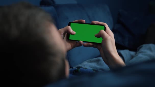 男人躺在沙发上，晚上用带有彩色键的绿色屏幕的智能手机，在社交媒体或网上商店里打转- -互联网，交流观念紧密相连. — 图库视频影像
