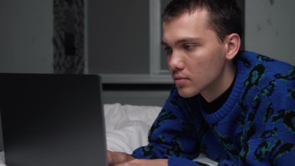 Junger gutaussehender kaukasischer Mann, der am Laptop arbeitet und zu Hause im Bett liegt. Mann surft im Internet oder chattet mit Freunden online auf Laptop entspannt im Bett zu Hause. — Stockvideo