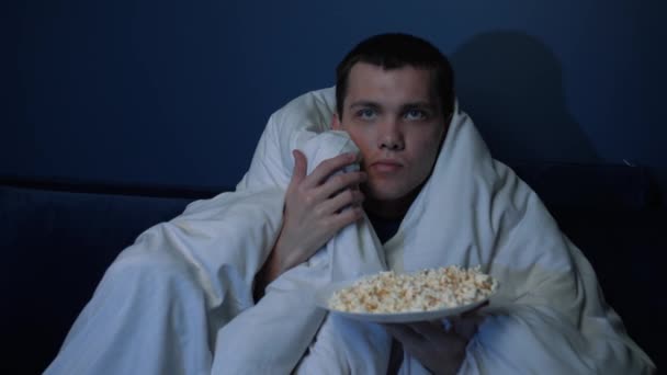 Bang knappe blanke man kijken horror film 's avonds laat, eten popcorn met deken, man kijken horror film op de bank thuis. — Stockvideo