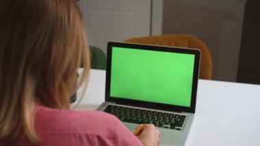 Yeşil ekran dizüstü bilgisayar konsepti: online uzaklık eğitimi, video konferansı. Ev çalışmasında omuz üstü çalışan bayan öğrenci bir not defterine bilgi yazar..