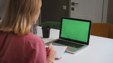 Yeşil ekran dizüstü bilgisayar konsepti: online uzaklık eğitimi, video konferansı. Ev çalışmasında omuz üstü çalışan bayan öğrenci bir not defterine bilgi yazar..
