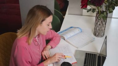 Kulaklıklı bir kadın dizüstü bilgisayarla çalışıyor, uzaktan eğitim alıyor, video konferansı düzenliyor. Evde çalışan bir kadın öğrenci not defterine bilgi yazar..