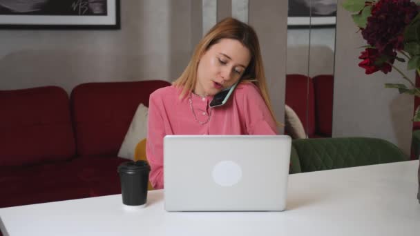 Güzel, genç bir iş kadını telefonda konuşuyor, bilgisayarını kullanıyor, ofis masasında oturuyor, mutlu bir kadın dizüstü bilgisayarla çalışıyor, ev ofisini arıyor.. — Stok video