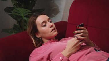 Rahatlamış Çekici genç kadın evdeki koltukta sosyal medyada online alışveriş için telefon kullanıyor, mobil mesaj iletişimi yavaş çekim