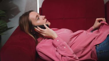 Evde ağır çekimde konuşan rahat bir kadın. Gülümseyen kız evdeki koltukta akıllı telefondan konuşuyor..