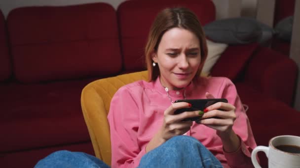 Νεαρή γυναίκα που παίζει κινητό παιχνίδι στο κινητό στο σπίτι. Ελκυστική γυναίκα απολαμβάνοντας smartphone βιντεοπαιχνίδια κάθεται στον καναπέ στο σαλόνι. — Αρχείο Βίντεο