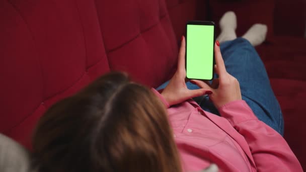 In Großaufnahme halten Frauenhände ein Smartphone mit grünem Chromakey-Bildschirm, das Inhalte anschaut, und wischen auf dem heimischen Sofa herum. Gadgets Menschen Konzept. — Stockvideo