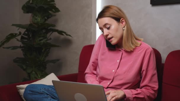 Druk bezette blanke vrouw zittend op de bank in de woonkamer, pratend op mobiele telefoon en werkend op laptop computer. Zakenvrouw aan de telefoon thuis. Vrouwelijke freelancer. — Stockvideo