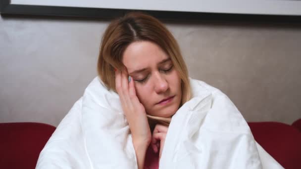 Joven chica caucásica enferma con covid o gripe sentado debajo de la manta en el sofá en la sala de estar y tienen dolor de cabeza. Mujer enferma con síntomas de coronavirus. — Vídeos de Stock