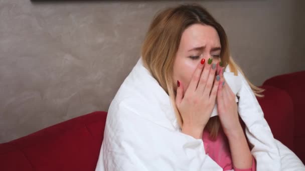 Joven chica caucásica enferma con covid o gripe sentado debajo de la manta en el sofá en la sala de estar y tos en servilleta. Mujer enferma con síntomas de coronavirus. — Vídeo de stock