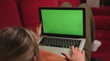 Yeşil ekranlı dizüstü bilgisayar kullanan genç bir kadın evdeki kanepede yatıyor. Arkadan bak. Krom anahtar