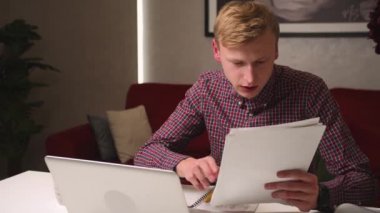 Ciddi genç işadamının web kamerasından konferans videosu var. Evraklarla çalışıyor, online öğretmenle çevrimiçi öğrenci çalışmalarına odaklanıyor. Evde dizüstü bilgisayarda görüntüler var. Adamın video konferansı var.