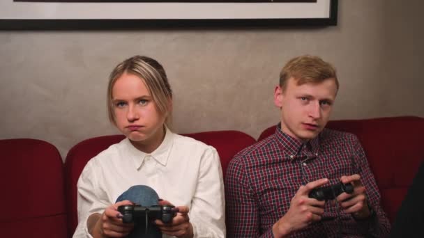 Молодые люди играют в видеоигры на консоли и скучно, зевая, сидя на диване перед телевизором. Тысячелетняя пара проводит время вместе дома — стоковое видео