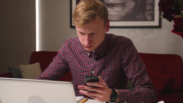 Hombre caucásico guapo usando el ordenador portátil escribiendo en el teléfono, trabajando en el escritorio de la oficina en casa. Hombre enfocado sosteniendo el teléfono. — Vídeos de Stock