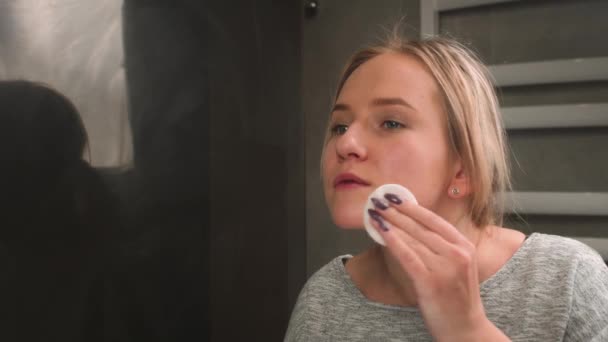 Mooie vrouw wast make-up af. Een vrouw in de badkamer kijkt in de spiegel en veegt make-up van haar gezicht met een wattenschijfje. Schoonheidsspecialiste, make-up alleen thuis concept. Close-up — Stockvideo