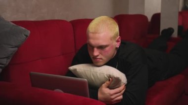 Yakışıklı homoseksüel adam kanepede dizüstü bilgisayar kullanarak yatıyor, rahat genç adam dizüstü bilgisayarında yazıyor, internette geziniyor, evde dizüstü bilgisayardan içeriği izliyor..