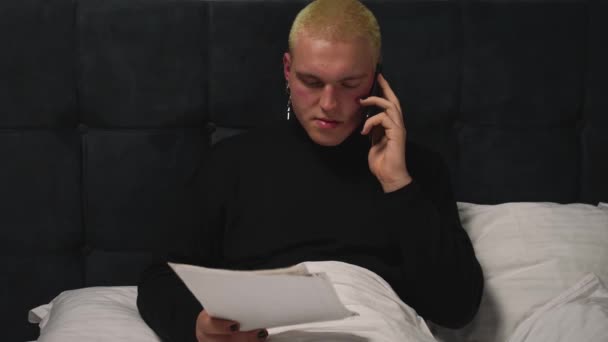 Przystojny homoseksualista pracujący z dokumentami na łóżku, rozmawiający przez telefon przed pójściem spać do domu. Mężczyzna z manicure pracuje z papierami na sypialni. — Wideo stockowe