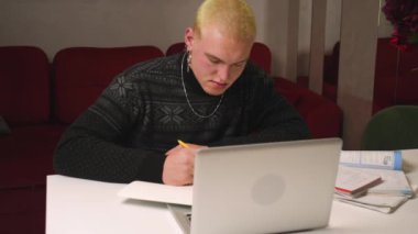 Dizüstü bilgisayar kullanan yorgun homoseksüel erkek öğrenci ödevlerini not defterine, evden online işe, internetten mesafelere yazıyor..